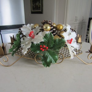 May include: A gold metal candelabra with five candle holders, decorated with a Christmas centrepiece featuring white poinsettias, green holly leaves, pine cones, and gold baubles.