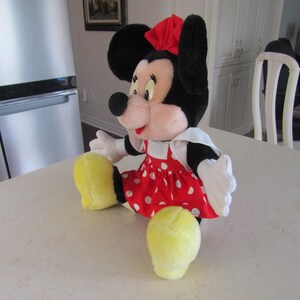 May include: A plush Minnie Mouse doll, featuring black ears, a red bow, and a red dress with white polka dots. The doll has white gloves and yellow shoes. The doll is sitting on a white surface.