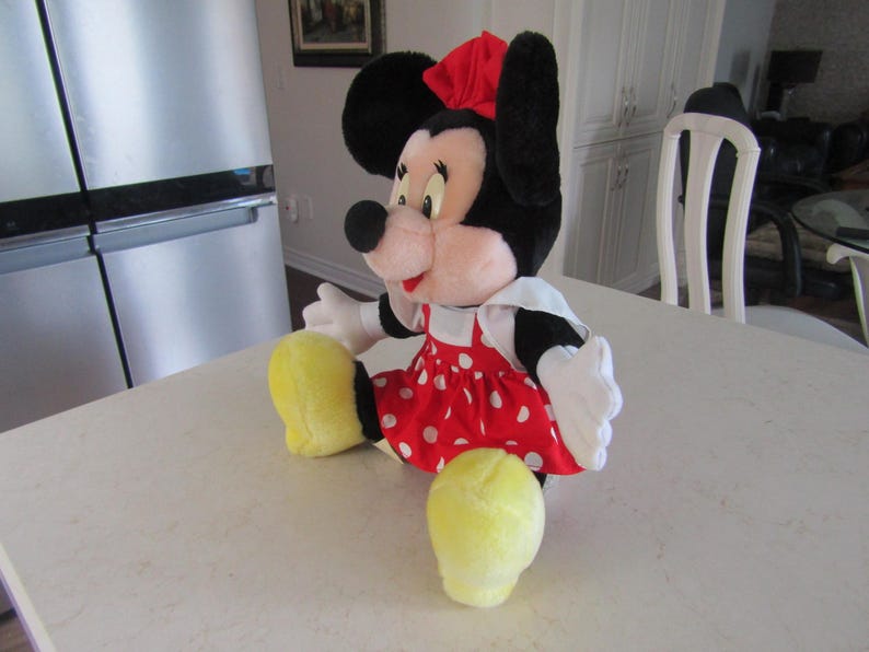 May include: A plush Minnie Mouse doll, featuring black ears, a red bow, and a red dress with white polka dots. The doll has white gloves and yellow shoes. The doll is sitting on a white surface.