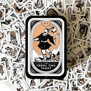 May include: A collection of The Teeny Tiny Tarot cards, featuring black and white illustrations with tan accents. The central card displays a figure with a hat, and the deck is surrounded by other cards with various scenes. The cards are arranged on a white surface.