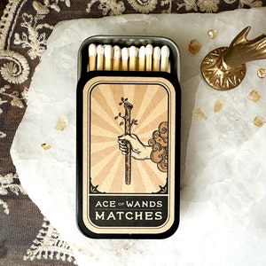 May include: A black and gold matchbox with a vintage illustration of a hand holding a wand with the text "Ace of Wands Matches".