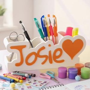 Könnte beinhalten: Ein weißer Schreibtisch-Organizer mit mehreren Fächern, der den Namen Josie in erhabenen orangefarbenen Buchstaben und ein orangefarbenes Herz zeigt. Der Organizer enthält verschiedene Stifte, Marker und eine Schere sowie weiteres Bastelzubehör.