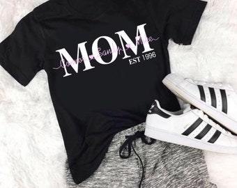 Può includere: Maglietta nera con testo bianco che dice "MOM" con i nomi "Connor, Bailey e Mike" scritti in corsivo sopra la parola "MOM". Il testo "EST 1996" è scritto sotto la parola "MOM".