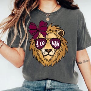 May include: Gray t-shirt featuring a lion graphic wearing sunglasses with the word "Lions" in a maroon font. The lion has a maroon bow on its head. The shirt is a casual style.