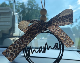 Peut inclure: Un charme de voiture noir avec le mot "mama" en écriture cursive. Le charme est suspendu à un rétroviseur avec un ruban à imprimé léopard et des perles en bois.