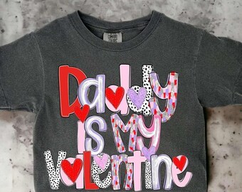 May include: Dark gray Comfort Colors t-shirt with the text "Daddy is my Valentine" in red, pink, purple, and white. The letters are decorated with hearts and patterns.