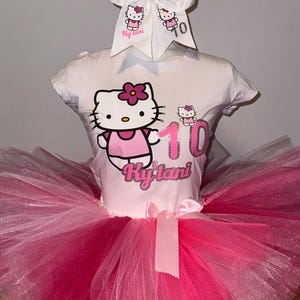 May include: A white t-shirt with a pink Hello Kitty graphic and the number 10. The shirt is paired with a pink and white tulle tutu and a white bow with pink Hello Kitty graphics and the name "Ky'lani" written on it.