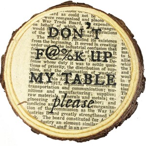 May include: A close-up of a wooden coaster with a printed text that reads "DON'T F@%K UP MY TABLE". The text is in black and the background is a light brown wood.