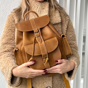 Könnte beinhalten: Ein brauner Lederrucksack mit Klappenverschluss, Schnalle und zwei Vordertaschen. Der Rucksack hat einen oberen Griff und verstellbare Träger. Die Tasche ist mittelgroß und scheint aus hochwertigem Leder zu sein.