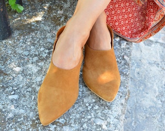 May include: A pair of brown suede pointed toe heeled mules. The shoes are worn by a person sitting on a stone surface.
