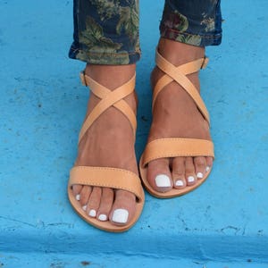 May include: Tan leather sandals with criss-cross straps and a flat sole. The sandals are worn on a person's feet with white nail polish.