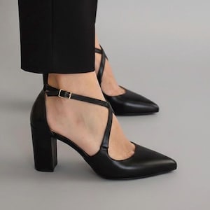 Peut inclure: Escarpins noirs en cuir avec bout pointu et talon bloc épais. Les chaussures présentent un design de lanières croisées et une bride de cheville avec une boucle dorée. Les talons mesurent environ 7.6 cm de haut.