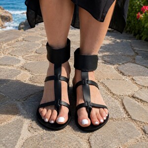 May include: A pair of black gladiator-style sandals featuring a wide ankle strap, a vertical T-strap design, and two horizontal straps across the foot. The sandals have a flat sole and a simple buckle closure system, providing a secure fit around the ankle.