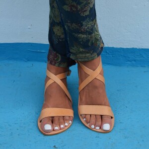 May include: Tan leather sandals with criss-cross straps and a buckle closure. The sandals have a flat sole and are worn on a person's feet.