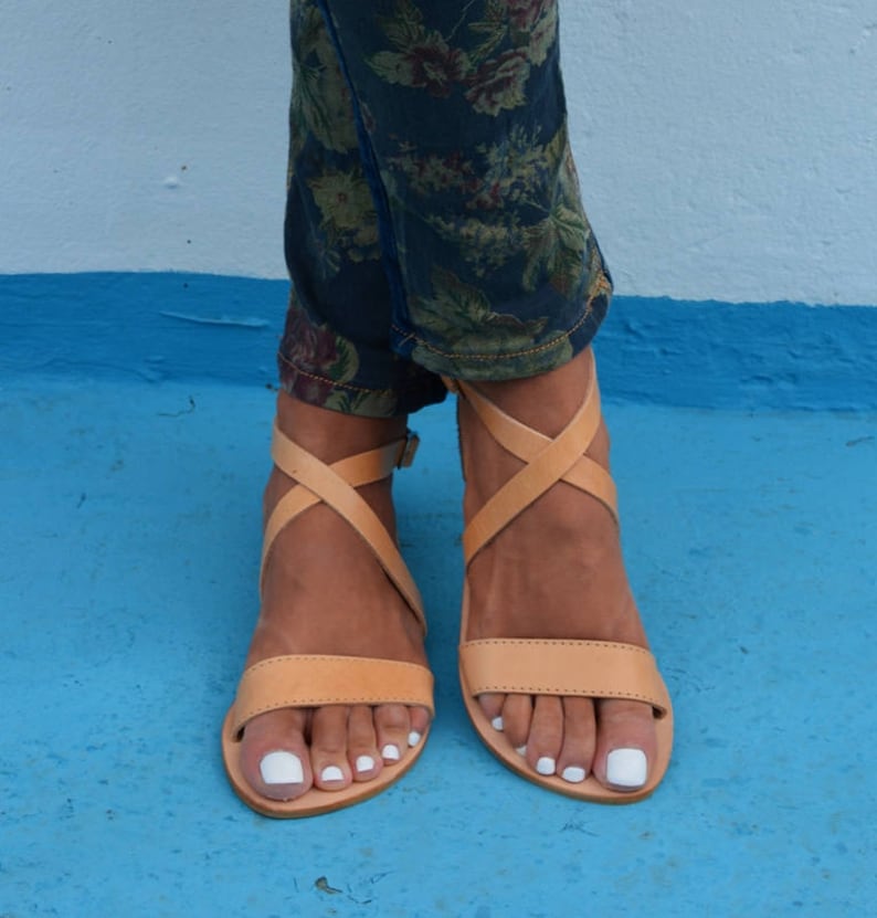 May include: Tan leather sandals with criss-cross straps and a buckle closure. The sandals have a flat sole and are worn on a person's feet.