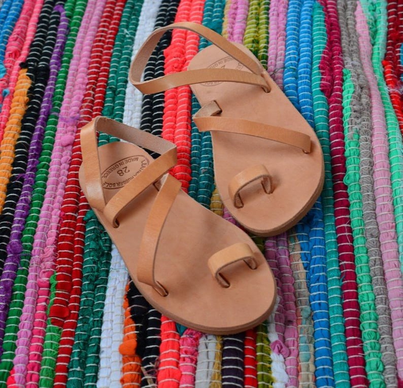 Pu&ograve; includere: Due paia di sandali in pelle color cuoio con pi&ugrave; cinturini. I sandali hanno un passante per le dita e sono realizzati in Grecia. I sandali sono su un tappeto a strisce colorate.