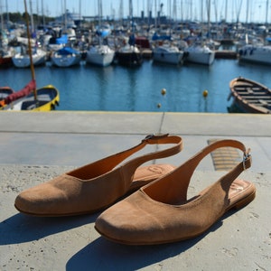 May include: Two pairs of light brown suede slip-on shoes with a slingback strap and buckle closure.