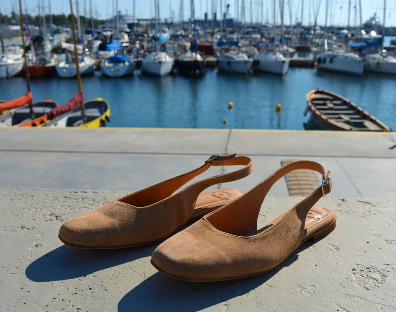 May include: Two pairs of light brown suede slip-on shoes with a slingback strap and buckle closure.