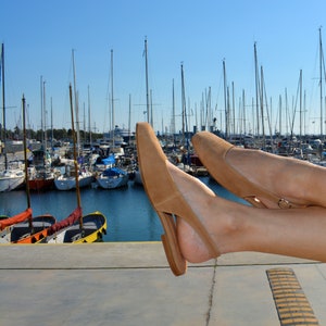 May include: A pair of tan suede heeled sandals with a slingback strap. The sandals are worn by a person's feet, with the toes of the sandals pointing towards the camera. The sandals are resting on a concrete surface with a yellow stripe.