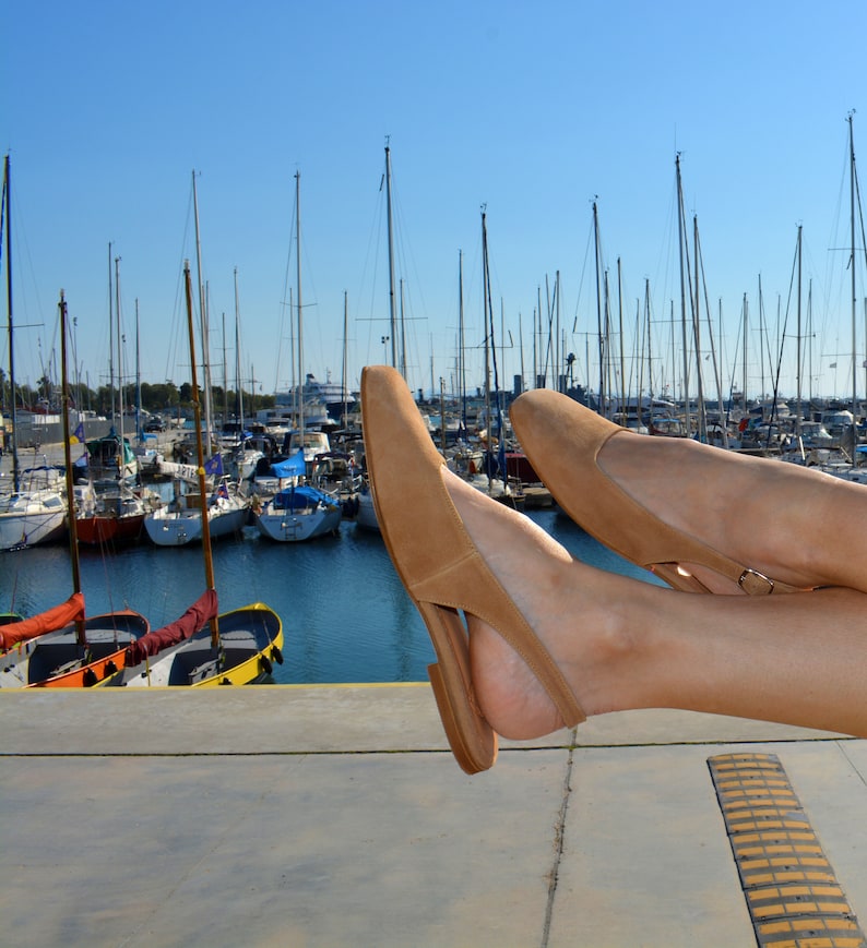 May include: A pair of tan suede heeled sandals with a slingback strap. The sandals are worn by a person's feet, with the toes of the sandals pointing towards the camera. The sandals are resting on a concrete surface with a yellow stripe.