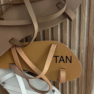 Puede incluir: Tres pares de sandalias de cuero en nude, marr&oacute;n claro y blanco. Las sandalias tienen una presilla para el dedo gordo y m&uacute;ltiples correas. La palabra "NUDE" est&aacute; impresa en la sandalia superior, "TAN" en la sandalia del medio y "WHITE" en la sandalia inferior.
