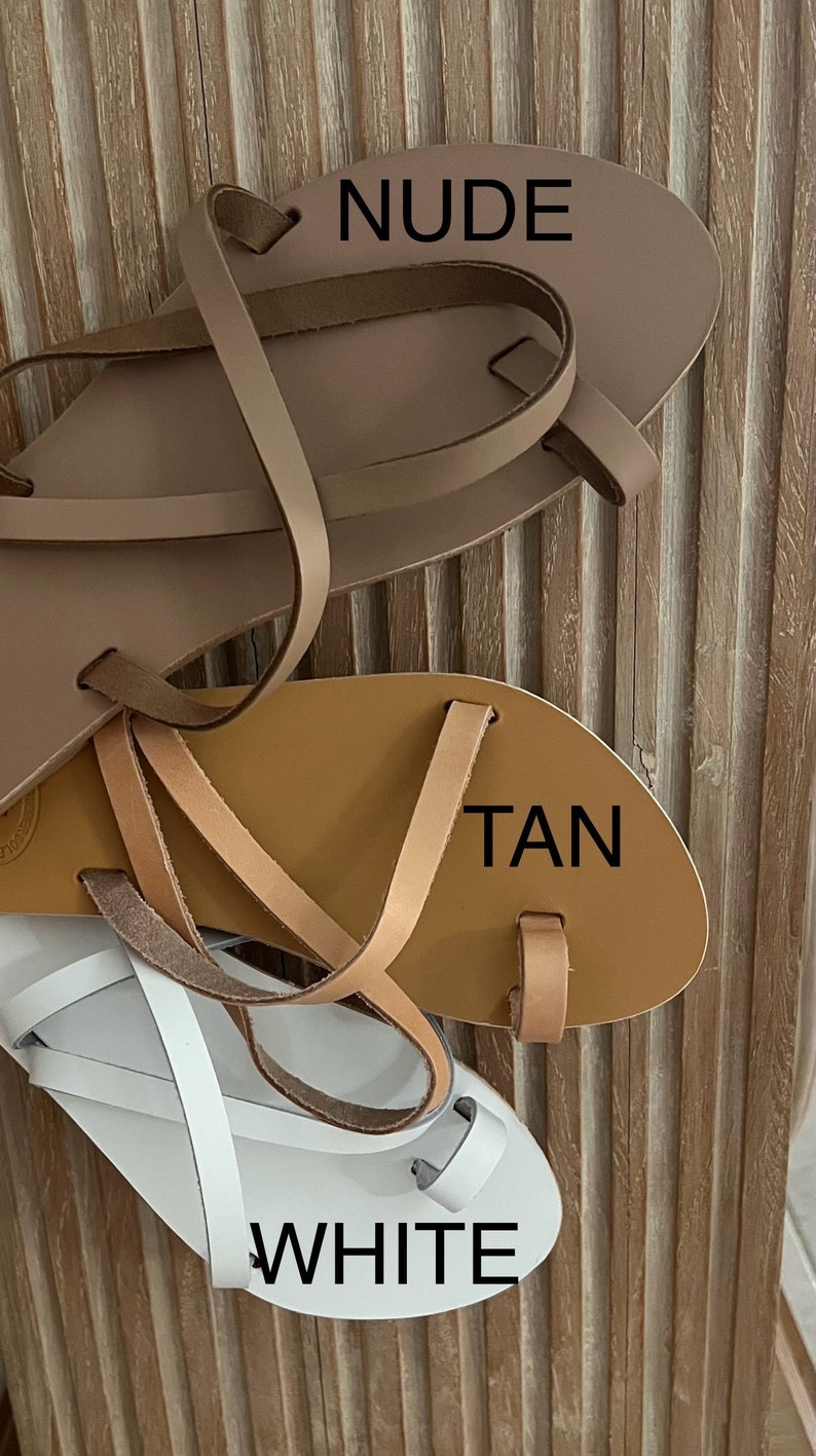 Puede incluir: Tres pares de sandalias de cuero en nude, marr&oacute;n claro y blanco. Las sandalias tienen una presilla para el dedo gordo y m&uacute;ltiples correas. La palabra "NUDE" est&aacute; impresa en la sandalia superior, "TAN" en la sandalia del medio y "WHITE" en la sandalia inferior.