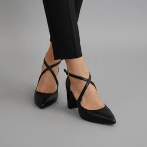 Op de afbeelding: Zwarte pumps met spitse neus, gekruiste bandjes en een blokhak. De schoenen hebben een enkelbandje met een gespsluiting. De hakken zijn ongeveer 7.6 cm hoog.