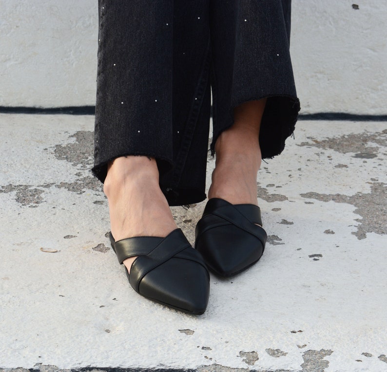 K&ouml;nnte beinhalten: Ein Paar schwarze, spitze Mules aus Leder mit einem gekreuzten Riemen-Design. Die Schuhe werden von einer Person getragen, die eine schwarze Jeans tr&auml;gt.
