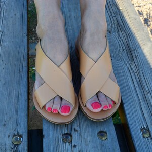 May include: A pair of light brown leather sandals with a crisscross strap design. The sandals are on a wooden surface. The image shows the sandals from a top-down perspective.