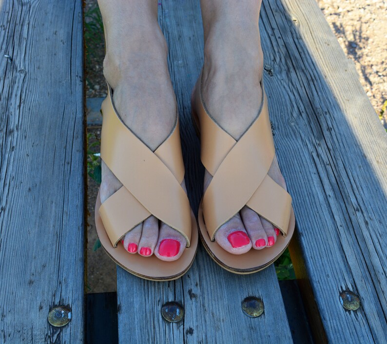 May include: A pair of light brown leather sandals with a crisscross strap design. The sandals are on a wooden surface. The image shows the sandals from a top-down perspective.