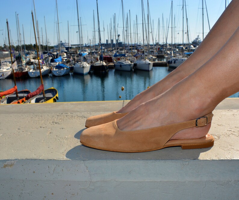 May include: Tan leather slip-on sandals with a single strap and buckle fastening. The sandals are on a concrete surface with a marina in the background.