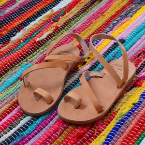 Pu&ograve; includere: Due paia di sandali in pelle color cuoio con cinturini e un passante per le dita. I sandali sono su un tappeto a strisce colorate.