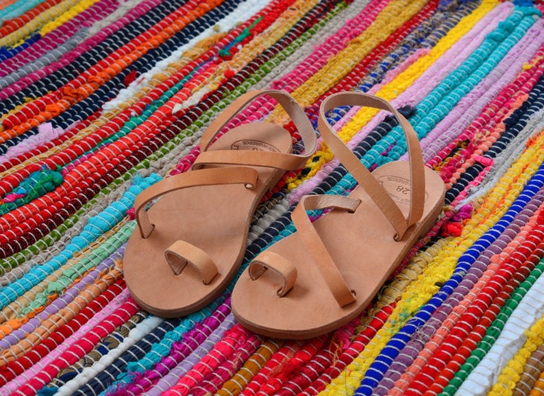 Pu&ograve; includere: Due paia di sandali in pelle color cuoio con cinturini e un passante per le dita. I sandali sono su un tappeto a strisce colorate.