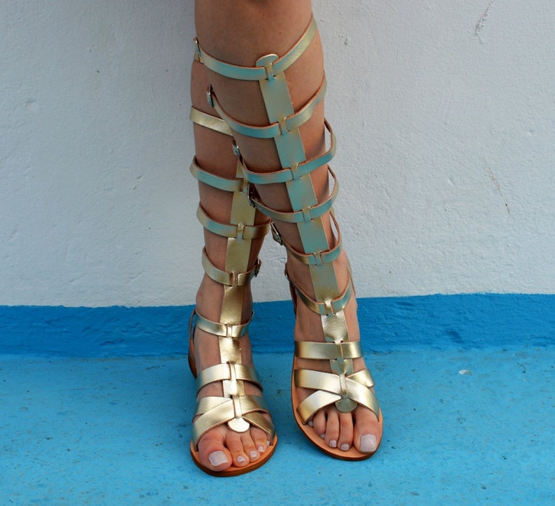 K&ouml;nnte beinhalten: Goldfarbene Gladiatorsandalen mit mehreren Riemen, die sich am Bein hochziehen. Die Sandalen haben eine flache Sohle. Sie werden vor einem wei&szlig;en und blauen Hintergrund getragen. Die Sandalen sind aus gl&auml;nzendem Material.