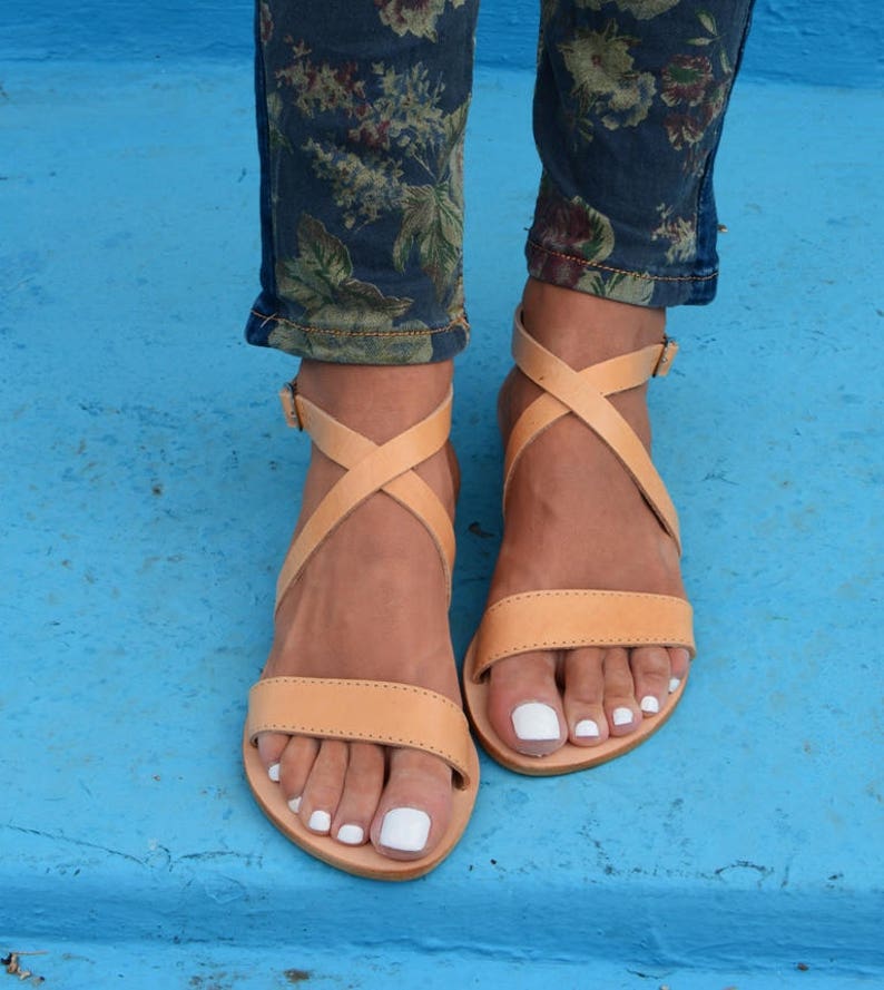May include: Tan leather sandals with criss-cross straps and a flat sole. The sandals are worn on a person's feet with white nail polish.