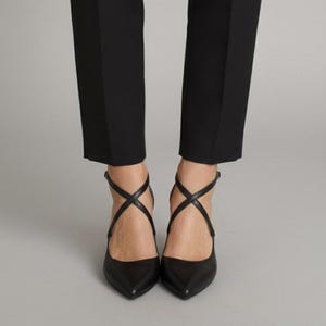 Op de afbeelding: Zwarte pumps met spitse neus en gekruiste enkelbandjes. De schoenen worden gecombineerd met een zwarte broek. De hakken zijn van een gematigde hoogte. Het ontwerp is strak en minimalistisch.