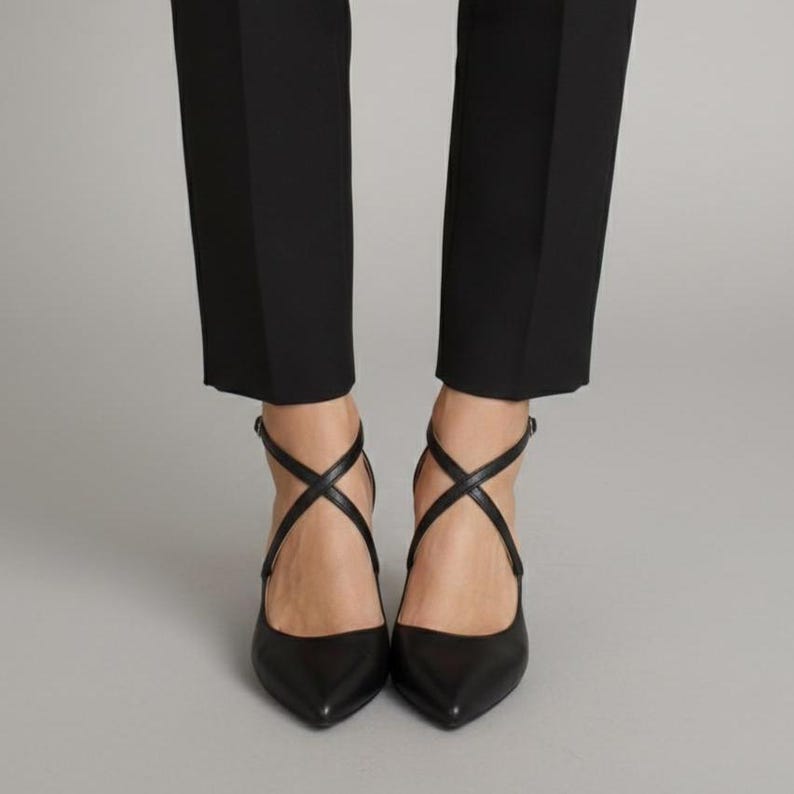Op de afbeelding: Zwarte pumps met spitse neus en gekruiste enkelbandjes. De schoenen worden gecombineerd met een zwarte broek. De hakken zijn van een gematigde hoogte. Het ontwerp is strak en minimalistisch.