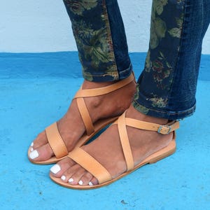 May include: Tan leather sandals with criss-cross straps and a buckle closure. The sandals have a flat sole and are worn on a person's feet.