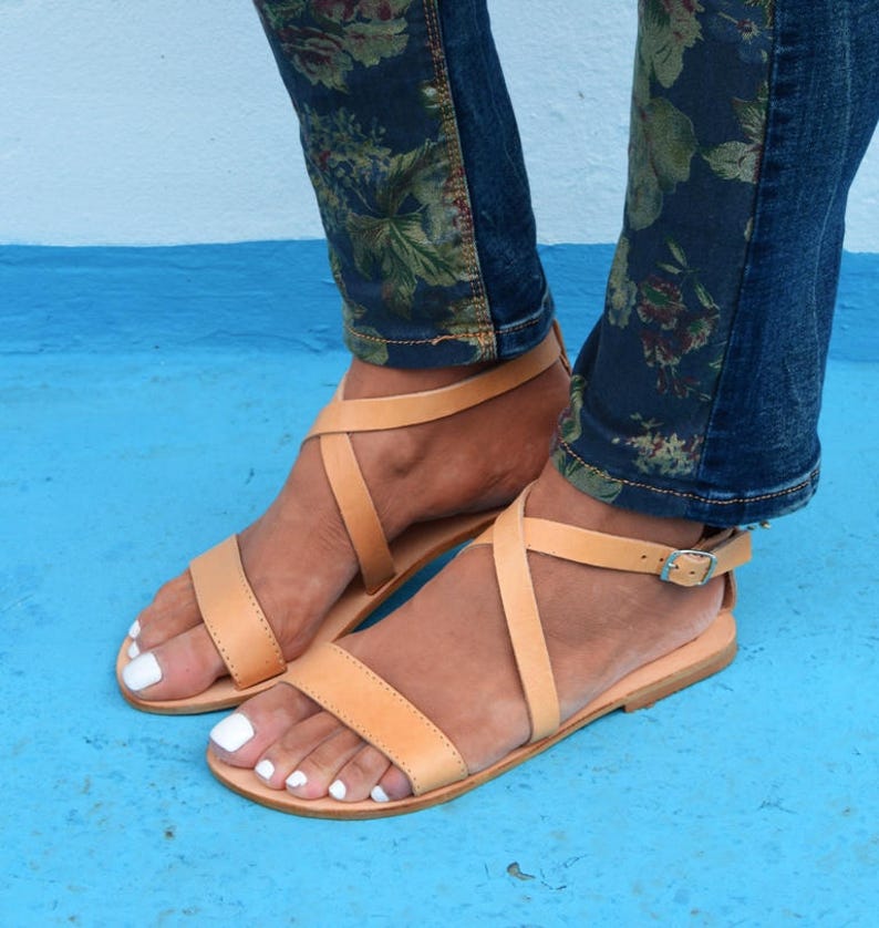 May include: Tan leather sandals with criss-cross straps and a buckle closure. The sandals have a flat sole and are worn on a person's feet.