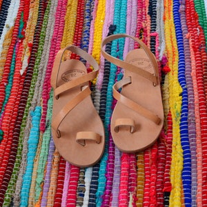 Pu&ograve; includere: Un paio di sandali in pelle color cuoio con un passante per l'alluce e pi&ugrave; cinturini. I sandali sono realizzati in Grecia e hanno la taglia 38 stampata sui cinturini.