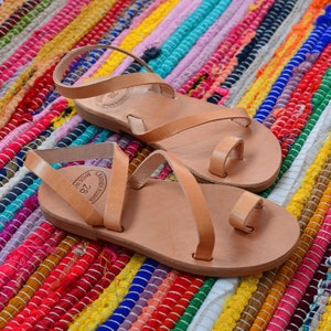 Pu&ograve; includere: Due paia di sandali in pelle color cuoio con cinturini incrociati e anelli per le dita. I sandali sono su un tappeto a strisce colorate.
