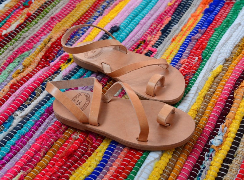 Pu&ograve; includere: Due paia di sandali in pelle color cuoio con cinturini incrociati e anelli per le dita. I sandali sono su un tappeto a strisce colorate.