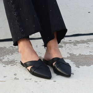 K&ouml;nnte beinhalten: Ein Paar schwarze Leder-Mules mit einem gekreuzten Riemen-Design und spitz zulaufenden Zehen.