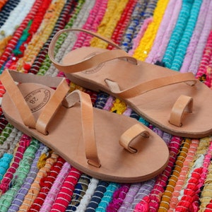 Pu&ograve; includere: Due paia di sandali in pelle color cuoio con un passante per le dita e un cinturino alla caviglia. I sandali sono su un tappeto intrecciato colorato.