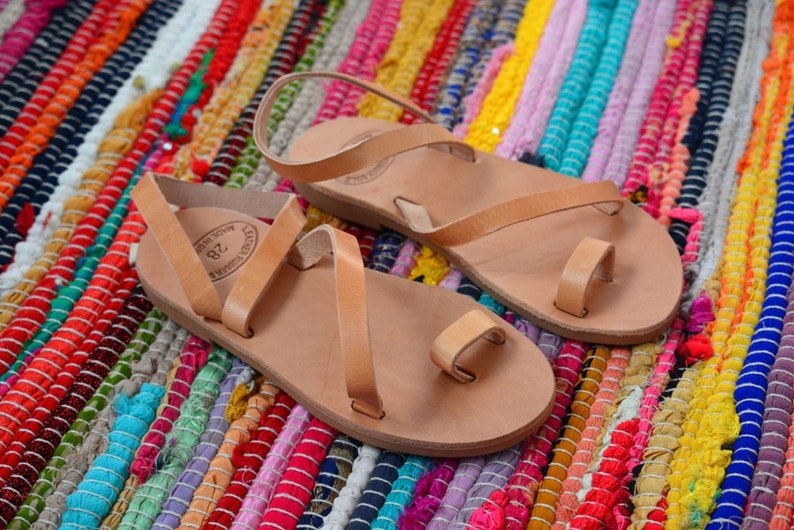 Pu&ograve; includere: Due paia di sandali in pelle color cuoio con un passante per le dita e un cinturino alla caviglia. I sandali sono su un tappeto intrecciato colorato.