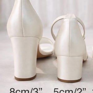 Op de afbeelding: Twee paar ivoorkleurige sandalen met blokhak. E&eacute;n paar heeft een hak van 8 cm (3"), de andere een hak van 5 cm (2"). De sandalen hebben een eenvoudig ontwerp met een enkelbandje.