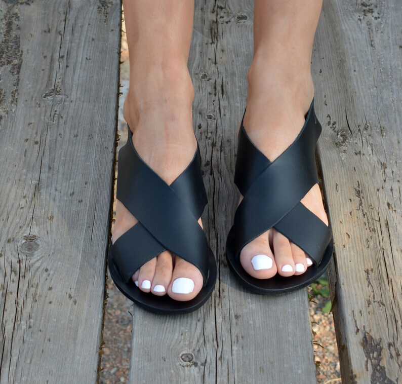 May include: Black leather sandals with a crisscross strap design. The sandals have a flat sole and are shown on a wooden surface. The sandals are a neutral color and can be worn with many outfits.