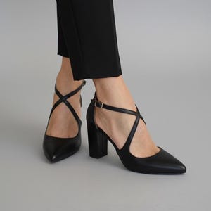 Op de afbeelding: Zwarte pumps met een spitse neus en gekruiste bandjes. De schoenen hebben een blokhak en een enkelbandje met een goudkleurige gesp. De schoenen worden gedragen met een zwarte broek.