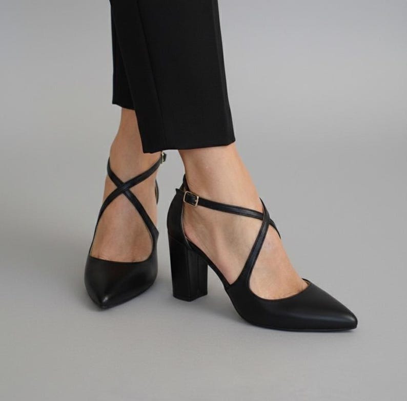Op de afbeelding: Zwarte pumps met een spitse neus en gekruiste bandjes. De schoenen hebben een blokhak en een enkelbandje met een goudkleurige gesp. De schoenen worden gedragen met een zwarte broek.