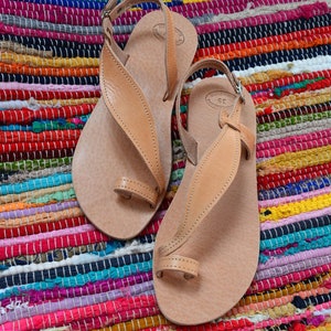 Puede incluir: Un par de sandalias de cuero marr&oacute;n claro con correas para los dedos y el tobillo. Las sandalias se exhiben sobre una colorida alfombra a rayas con tonos de rojo, rosa, azul y amarillo. Dise&ntilde;o minimalista.